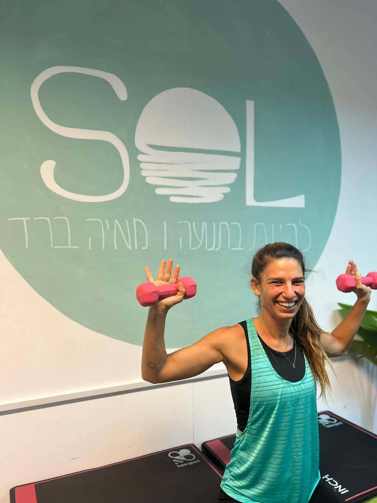 מאיה ברד עם משקולות יד מול קיר הלוגו של SOL